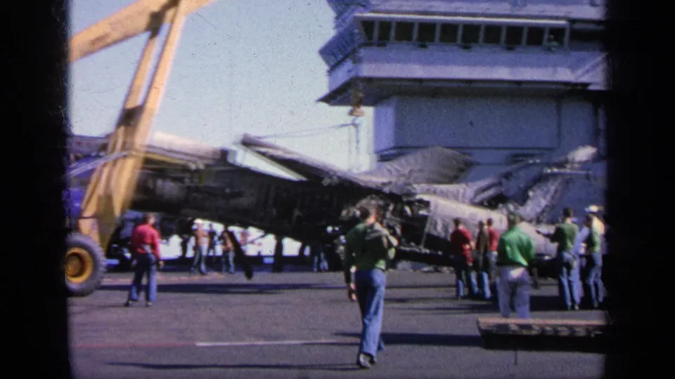 Deck-level 1963 Greece 8mm view of observers near a damaged military aircraft, a yellow crane, and the aircraft carrier island.