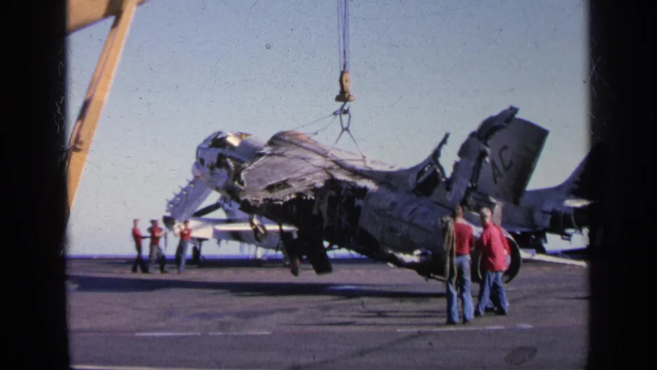 1963 Greece 8mm frame showing a crane lifting a badly damaged military aircraft while observers stand on an aircraft carrier deck.