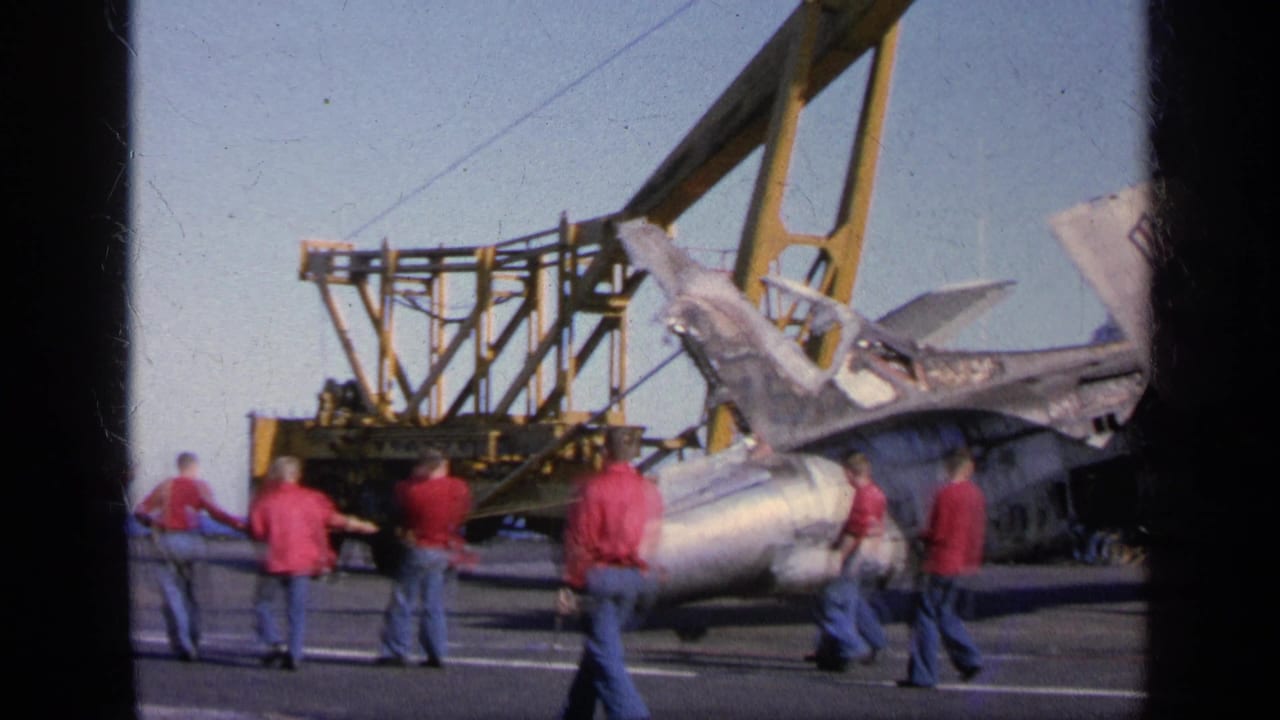 Deck crew guide a damaged military jet near a crane on a naval vessel deck.