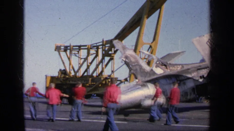 1963 8mm frame from Greece showing crew in red shirts guiding a damaged U.S.-marked military jet near a shipboard crane.
