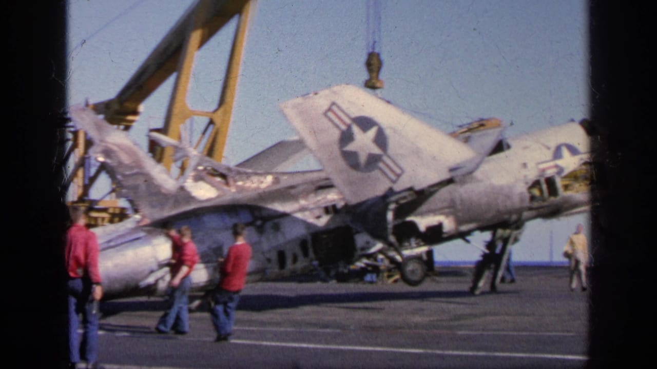 A damaged military aircraft is positioned under crane equipment on a ship deck.