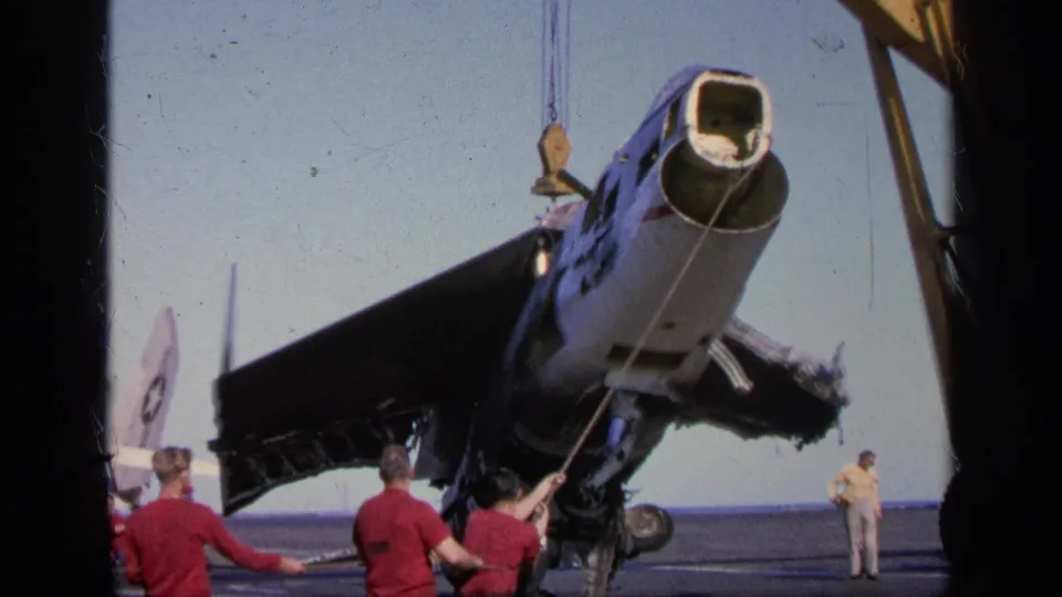 1963 Greece 8mm frame showing crew controlling lines as a damaged military aircraft is lifted or stabilized on a naval deck.
