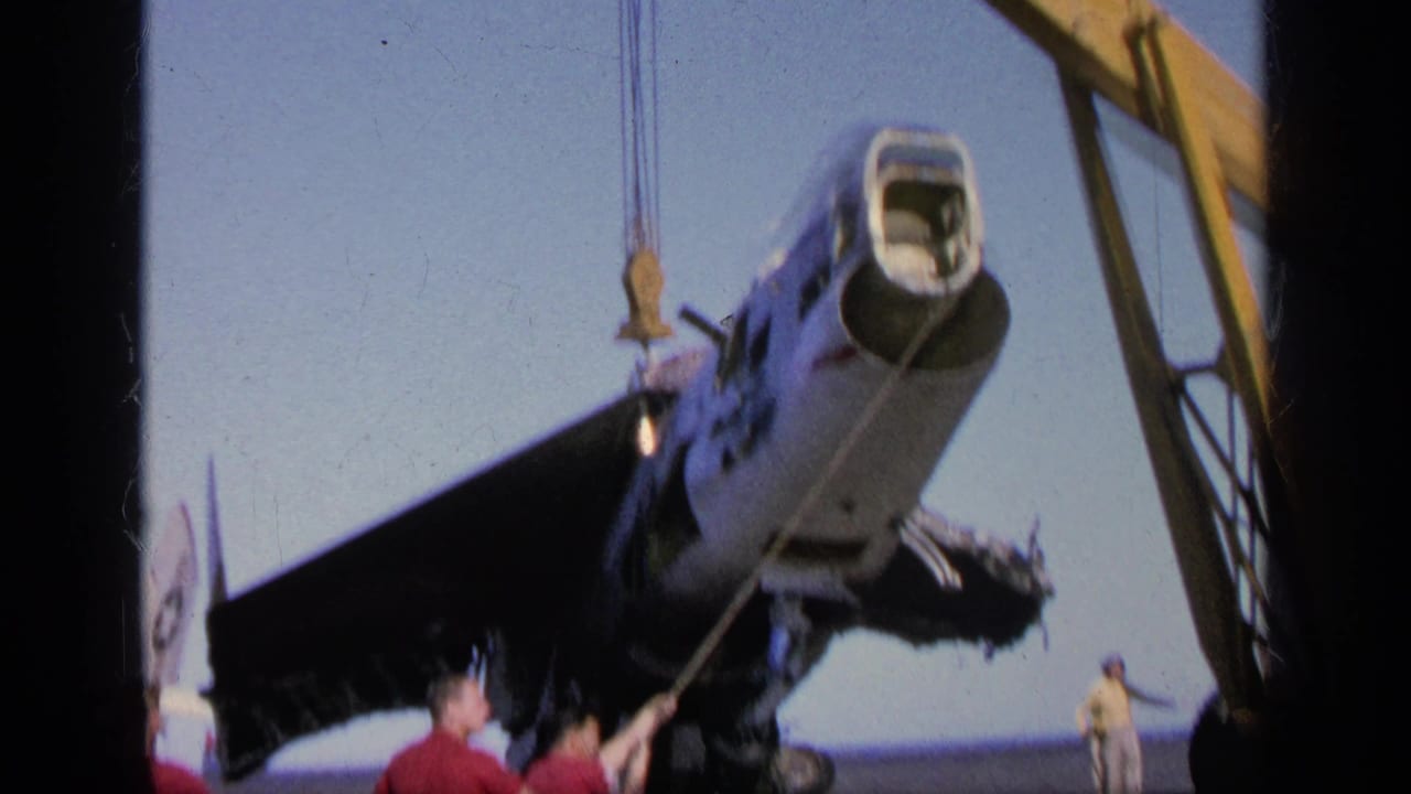 1963 home-movie frame near Greece showing sailors guiding a military jet suspended by crane over an aircraft carrier deck.