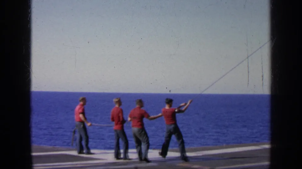 1963 Greece deck scene with sailors in red shirts pulling a rope to steady an aircraft off frame over the sea.