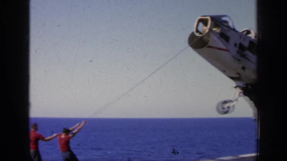 Home-movie frame from 1963 near Greece showing a military jet hanging above the deck while sailors control a guide rope.