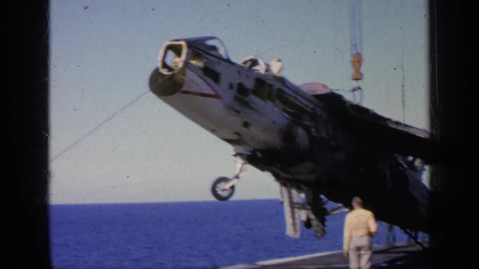 1963 Greece carrier-deck frame showing a military jet hoisted by crane, with a sailor nearby and ocean visible beyond.