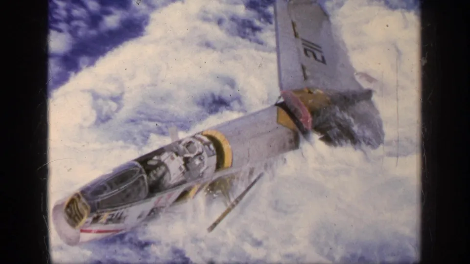 1963 Greece 8mm frame showing a marked military jet angled downward against bright clouds.