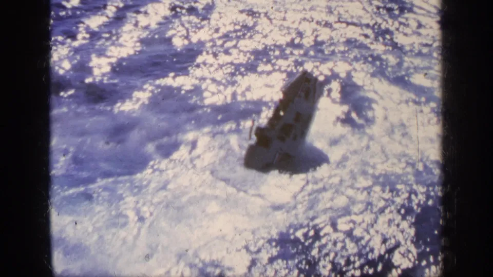 A marked aircraft section sits low in the sparkling ocean, appearing partly submerged after impact.