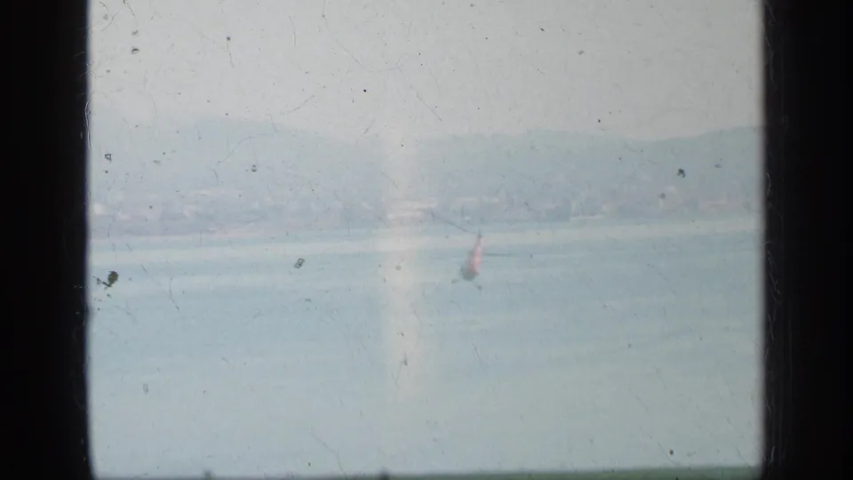 Hazy 1963 Greece home-movie frame looking across pale sea toward a distant shoreline, with film dust and a small red vertical feature near the water.