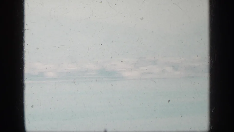 Soft 1963 8mm view of a hazy Greek coastal shoreline, pale water, low buildings, and mountains in the distance.