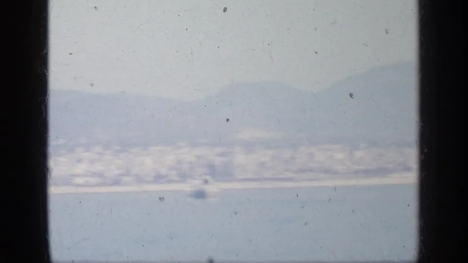 Hazy 8mm coastal view in Greece, with water in the foreground, distant buildings, mountains, and a small indistinct object near shore.