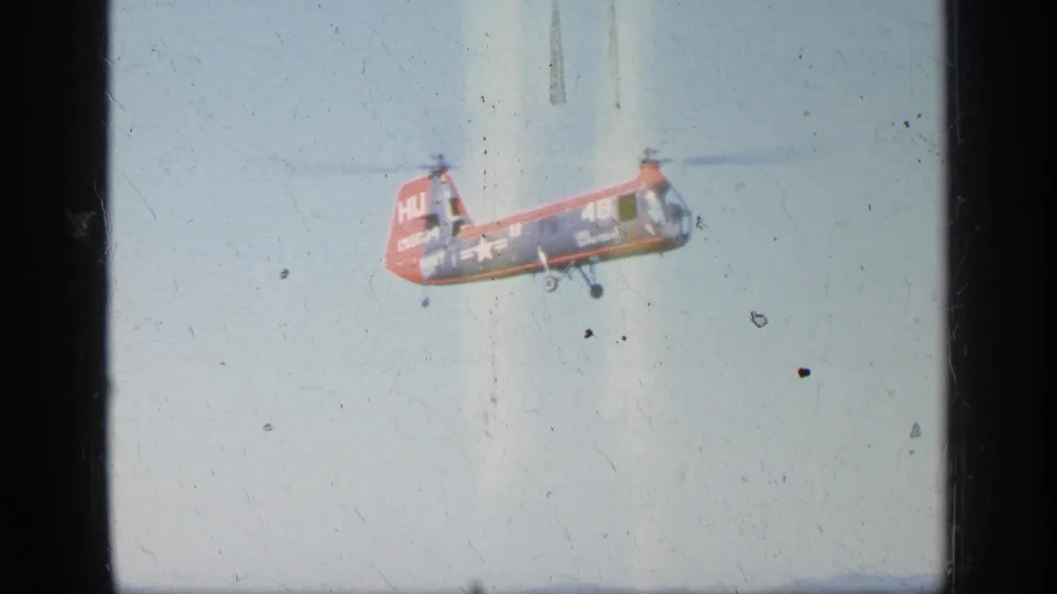 Red helicopter in flight against a pale sky, captured on grainy 1963 8mm home-movie film with visible dust and scratches.