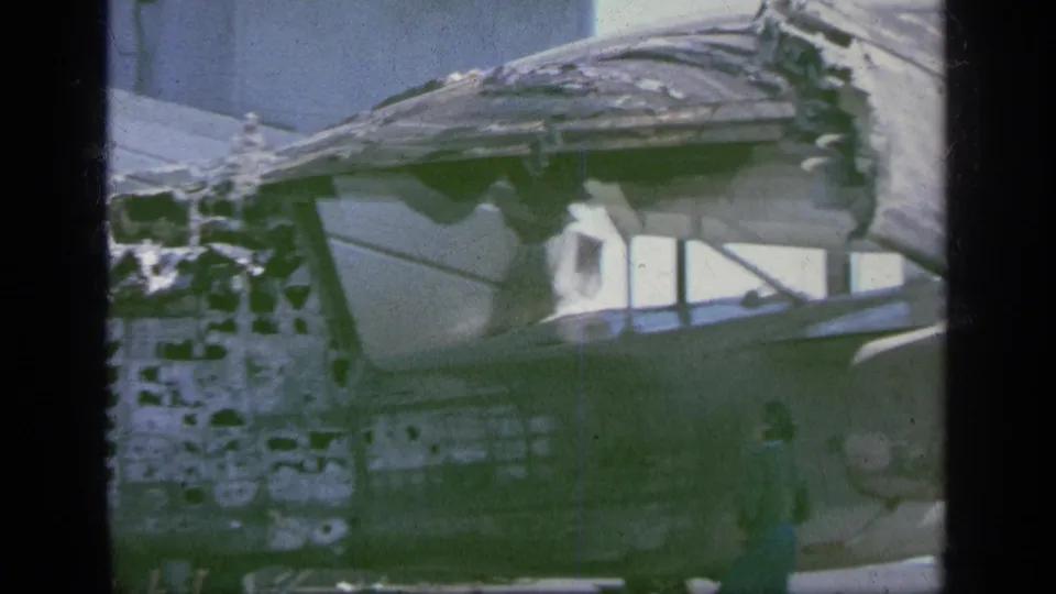 Weathered or damaged structure by the water, filmed in soft 8mm color with shoreline and pale sky beyond.