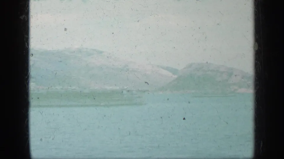Distant Greek shoreline and low mountains across hazy blue-green water in a faded 1963 8mm home-movie frame.