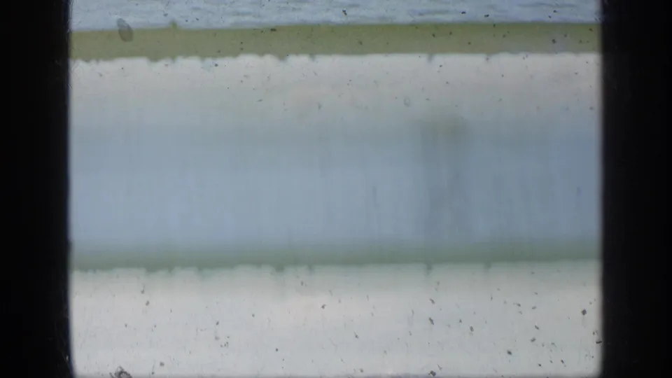 Soft 1963 Greece coastal frame with blurred water, a distant shoreline band, and pale sky, marked by dust and fading.
