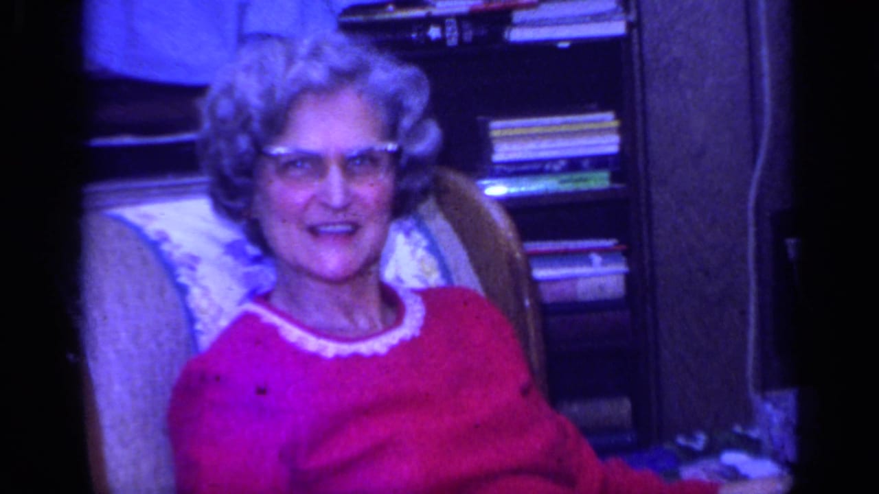 Elderly woman with glasses in a red sweater sits in a patterned armchair.