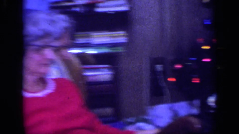 The handheld camera moves past the seated woman in the living room, creating motion blur and saturated 8mm color.