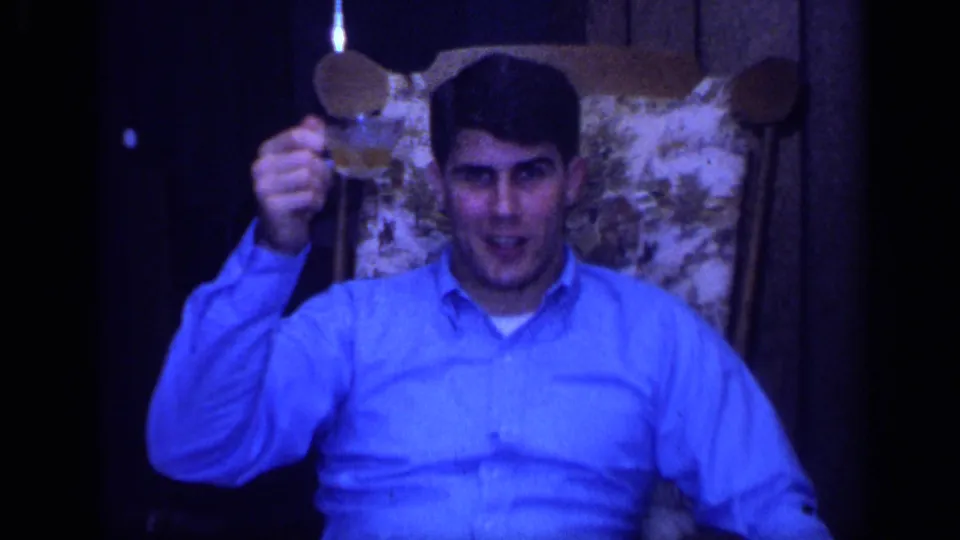 A young man in a blue shirt sits in a patterned chair and raises a small object toward the 8mm camera.