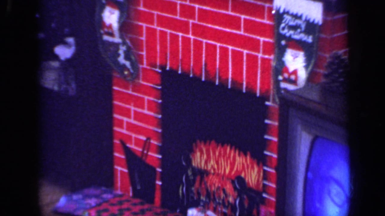 1968 California home-movie frame of a red brick fireplace display with Christmas stockings and mantel decorations.