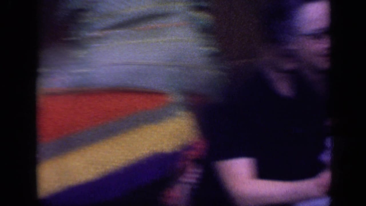 Blurred person in a dark shirt sits near colorful bedding or cushions.