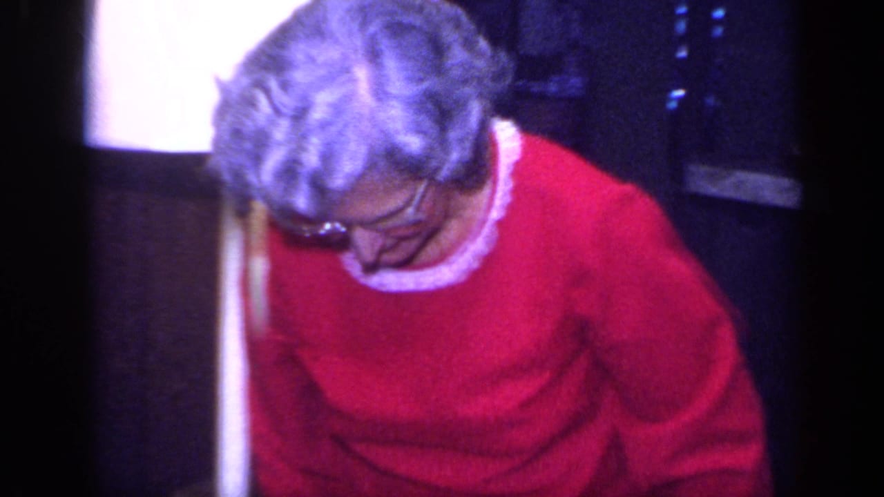 An older person wearing glasses and a red sweater looks downward in a softly exposed 1968 California home-movie interior.