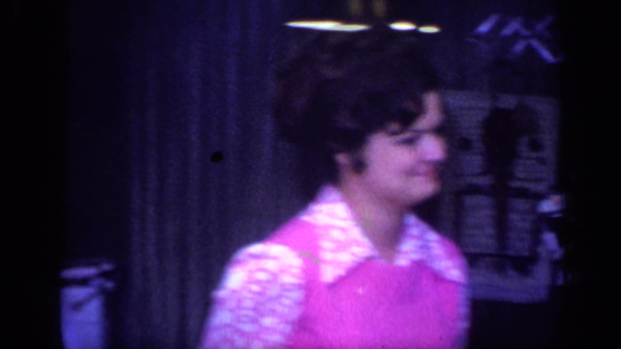 A woman in a pink dress with a patterned collar stands near a vintage camera in a 1968 California home-movie interior.