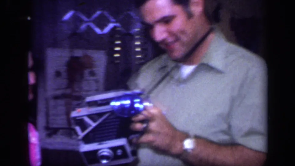 In supported 1968 California home-movie footage, a man smiles while holding an unidentified glowing blue object beside a partial woman indoors.
