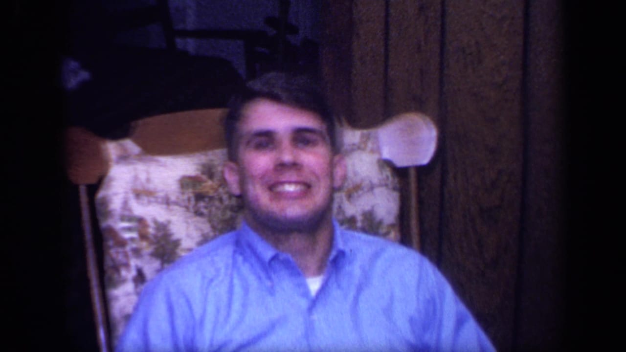 In the 1968 California home-movie setting, a smiling man in a blue shirt sits in a patterned chair in a wood-paneled room.