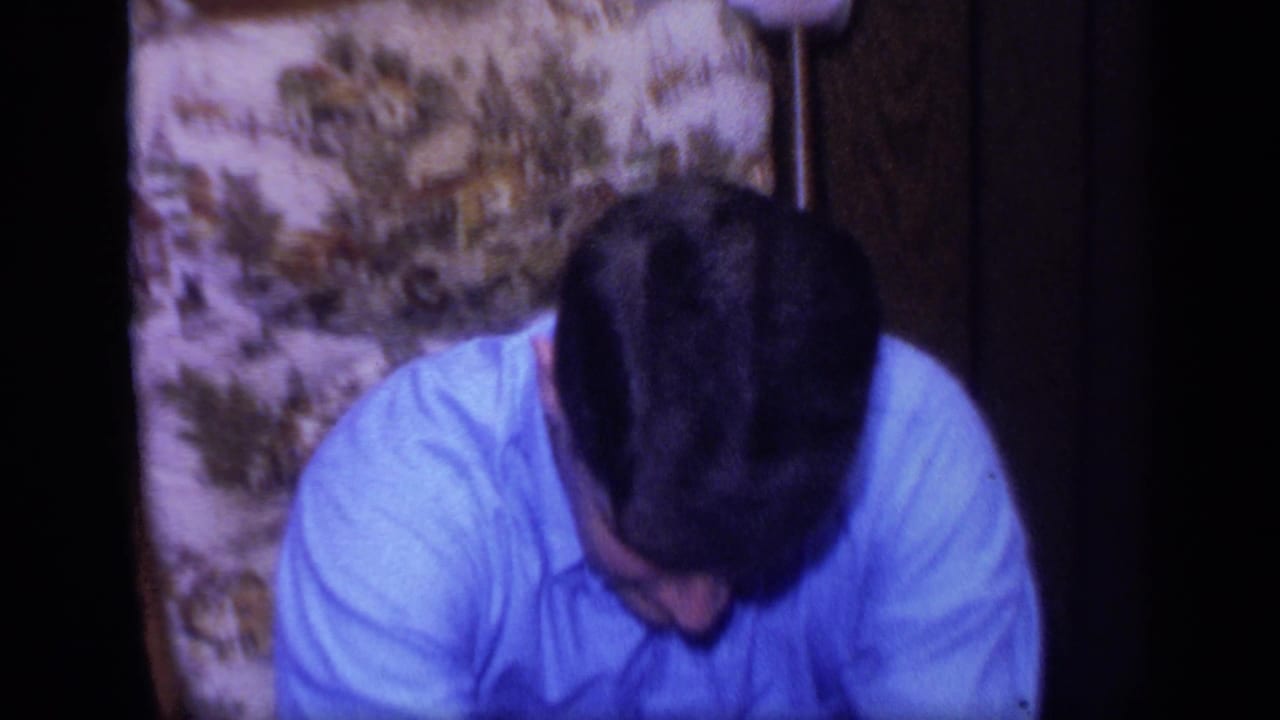 A seated man in a blue shirt looks downward in the same wood-paneled 1968 California home-movie interior.