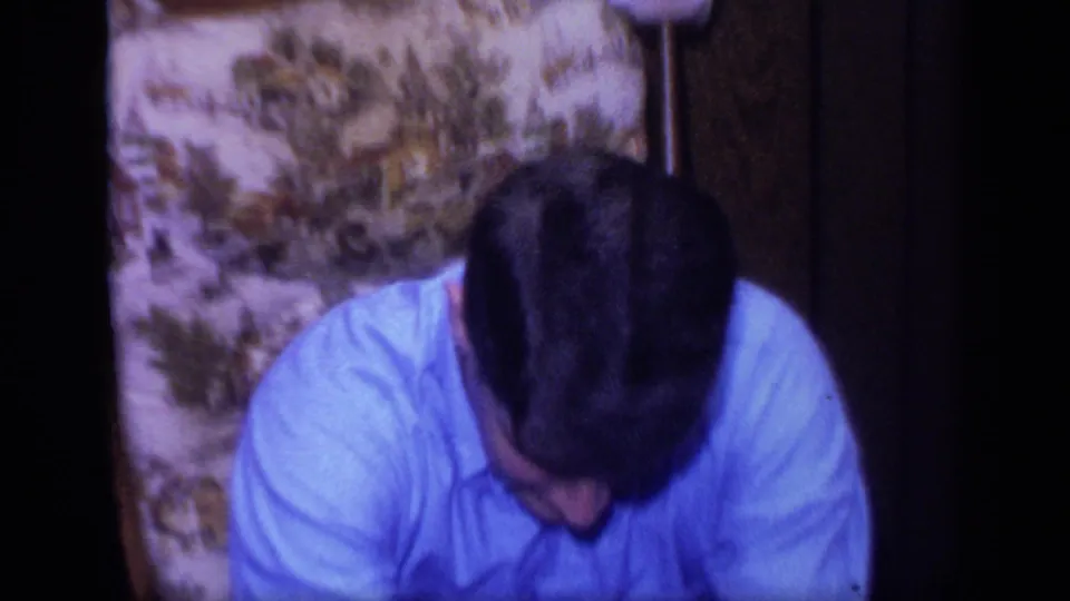 A seated man in a blue shirt looks downward in the same wood-paneled 1968 California home-movie interior.