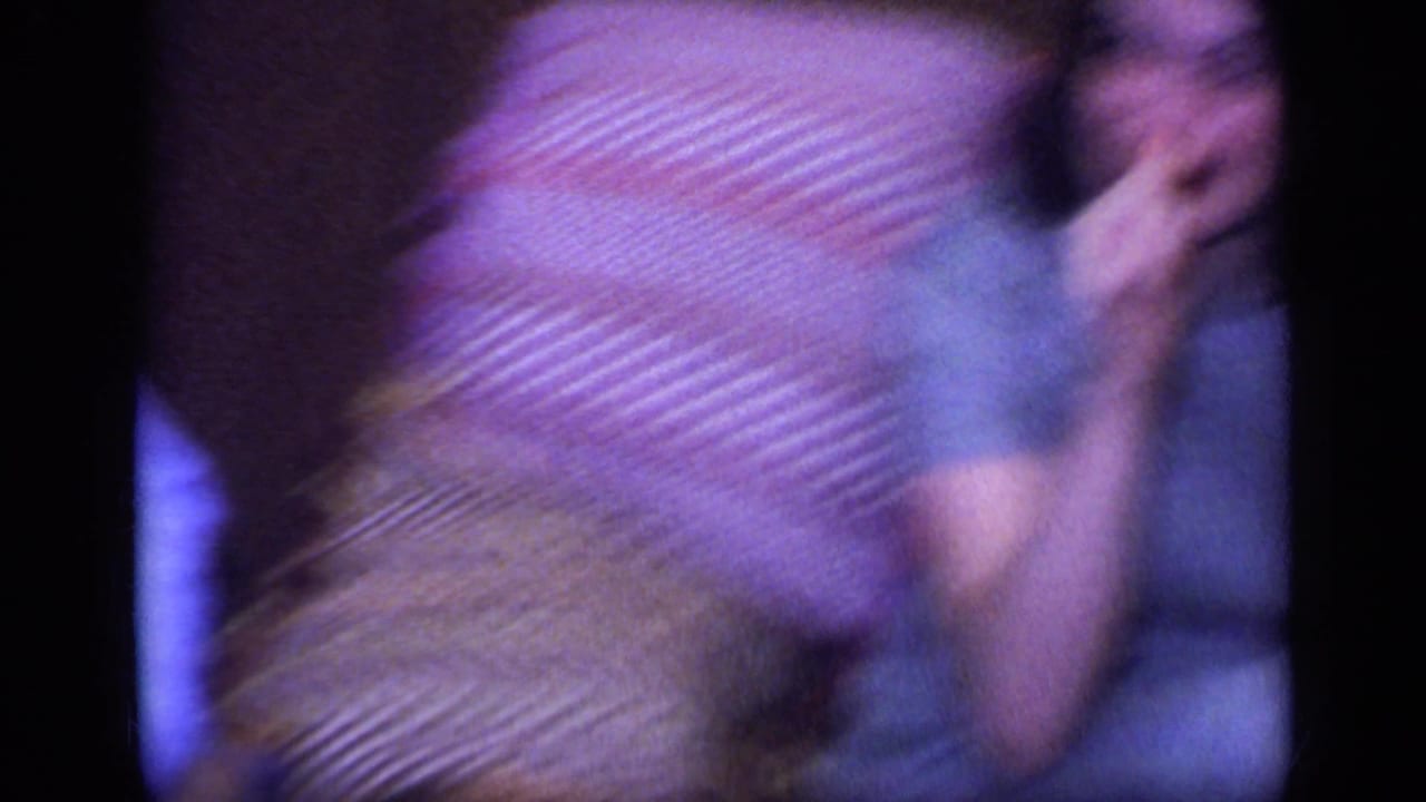 A motion-blurred 1968 California home-movie frame shows the seated woman and patterned sofa blanket during camera movement.