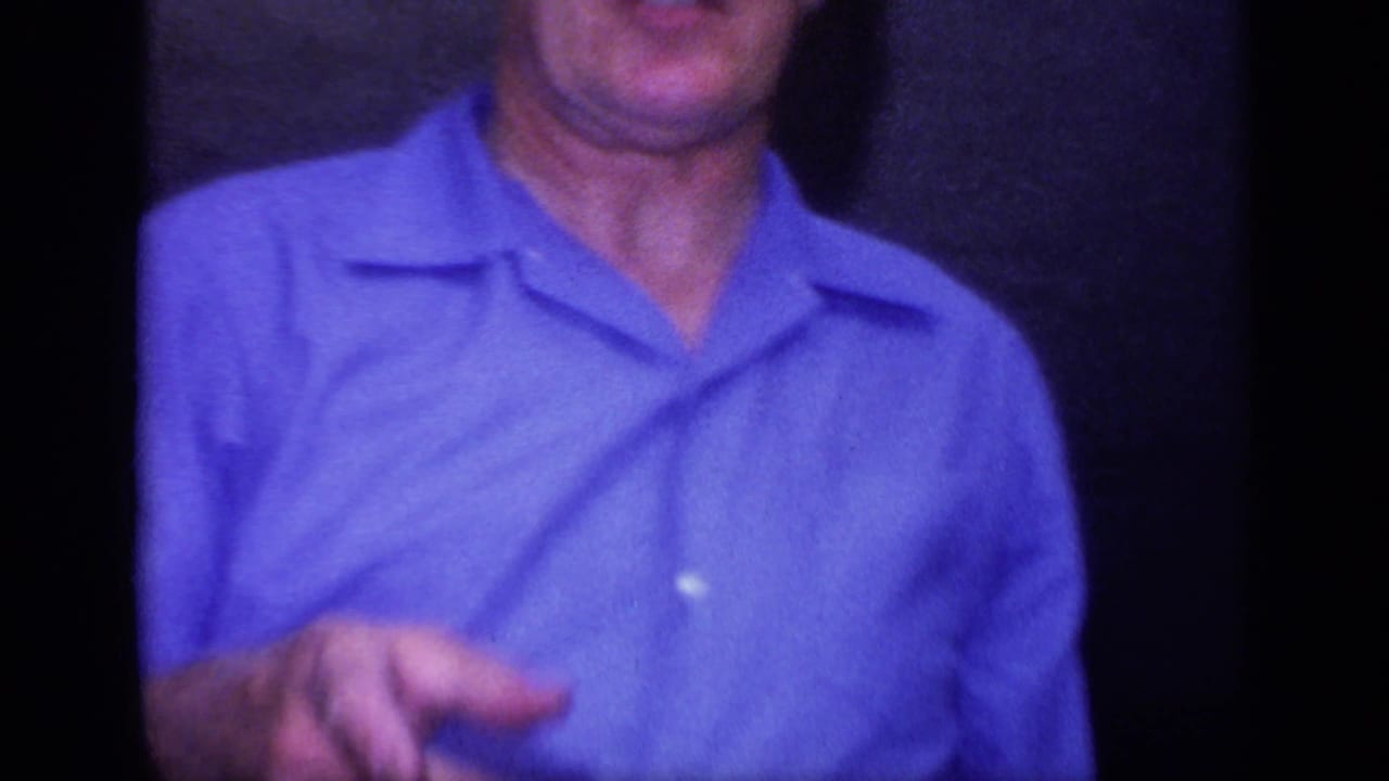 A close handheld frame shows an adult man in a blue collared shirt, cropped at the face and standing in a private interior.