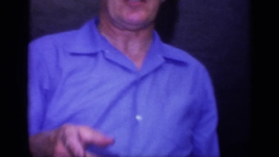 A close handheld frame shows an adult man in a blue collared shirt, cropped at the face and standing in a private interior.