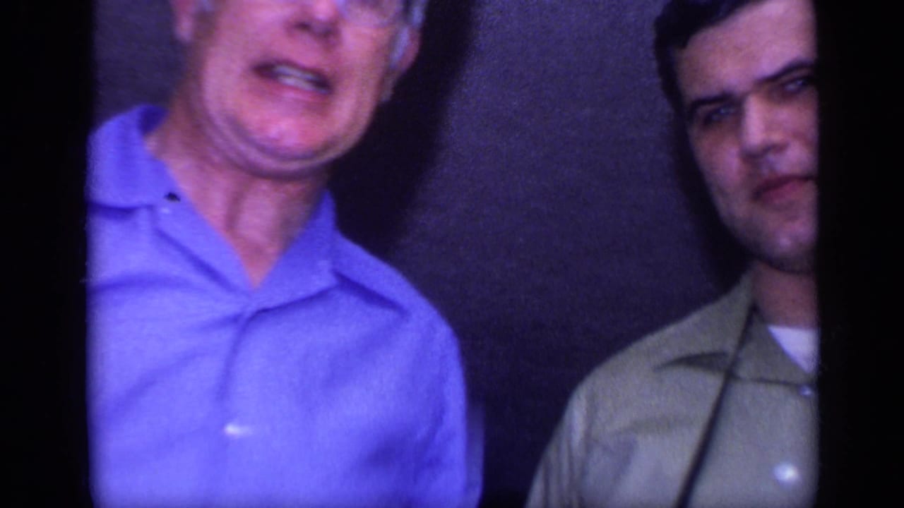 Two adult men stand indoors in a 1968 California home-movie frame, one in a blue shirt and one in a green collared shirt.