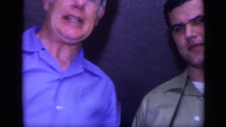 Two adult men stand indoors in a 1968 California home-movie frame, one in a blue shirt and one in a green collared shirt.