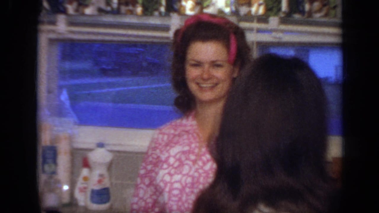 Smiling woman in a pink patterned top stands by a kitchen window as another woman faces away.