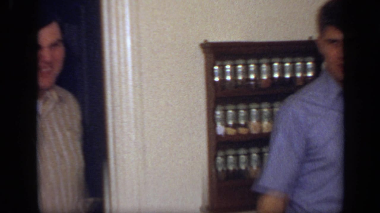 Smiling man in a striped shirt stands in a doorway while another man is partly visible near a spice rack.