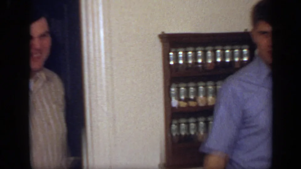 A smiling adult in a striped shirt appears in the doorway of a California kitchen, with another man partly framed beside a spice rack.