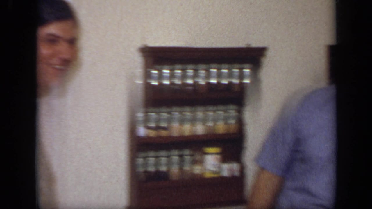 A smiling man is partly visible at the left edge as the handheld camera centers on a wooden spice rack in the 1968 kitchen.