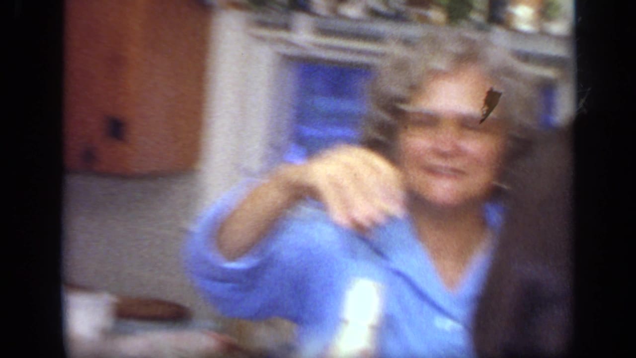 A woman with curly gray hair and glasses, wearing a blue shirt, smiles while holding a glass in the California kitchen.