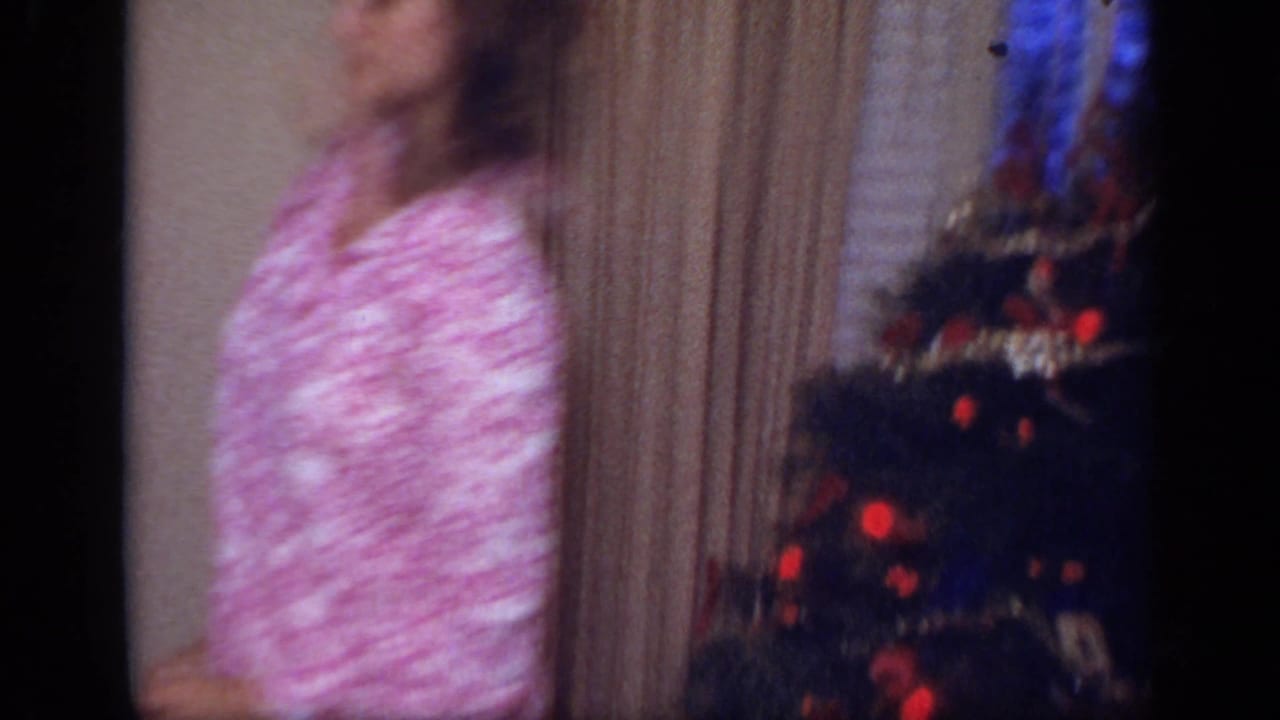 Woman in pink patterned pajamas stands near a decorated Christmas tree indoors.