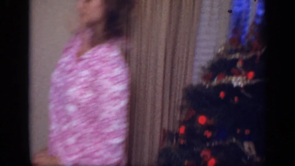 In a 1968 California home-movie frame, a woman in pink patterned pajamas stands beside a decorated Christmas tree in a private interior.