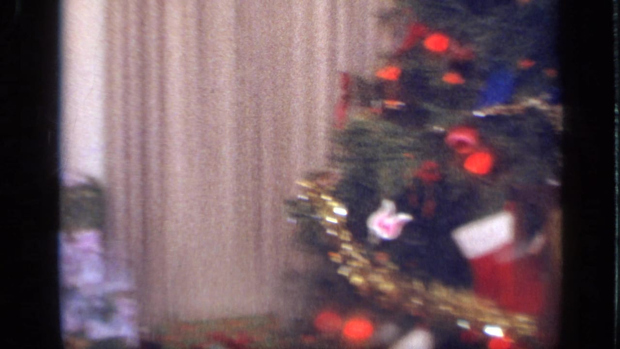 A decorated Christmas tree fills the frame beside curtains, with red ornaments, tinsel, and a stocking visible in the soft 8mm image.