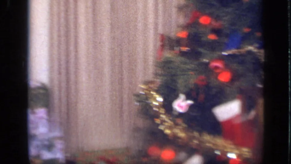 A decorated Christmas tree fills the frame beside curtains, with red ornaments, tinsel, and a stocking visible in the soft 8mm image.