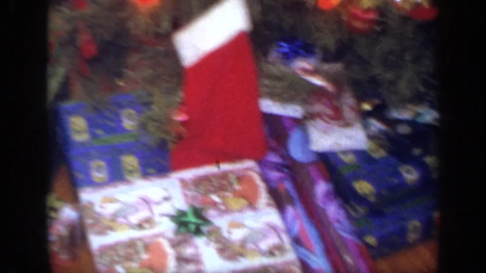 Wrapped presents, a red stocking, ornaments, and tinsel are gathered beneath the Christmas tree in the 1968 home-movie frame.