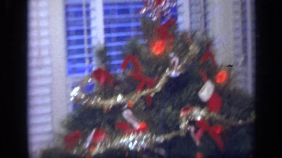 1968 home-movie frame of a Christmas tree with red ornaments and garland standing before interior window blinds.
