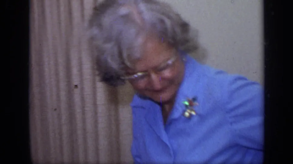 Older woman with gray hair, glasses, and a blue blouse sits near curtains in a 1968 California home-movie frame.