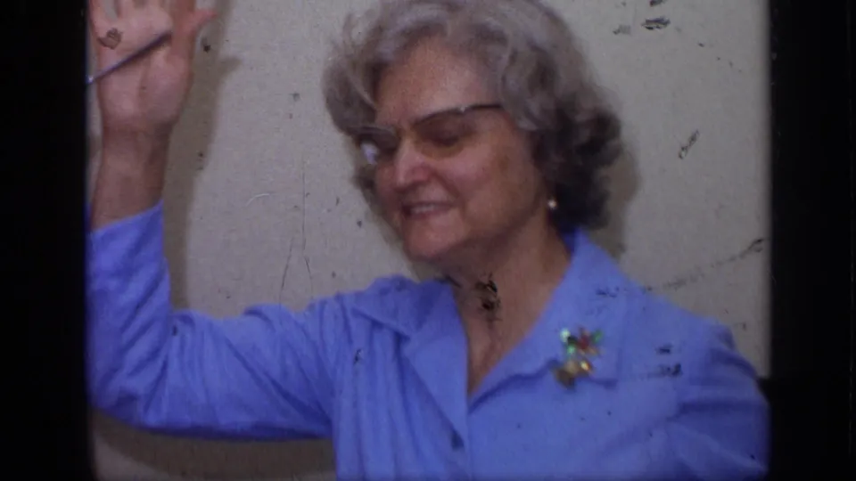 Older woman wearing glasses and a brooch smiles and waves in a 1968 California home-movie interior.