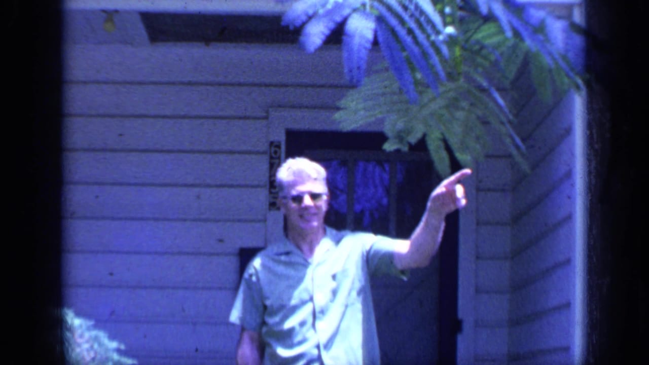 Man in sunglasses stands near a house doorway and points off-camera.