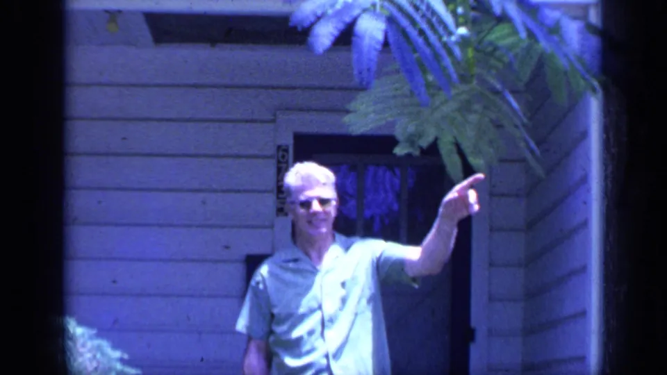 In 1968 Florida home-movie footage, a man in sunglasses stands by a house entrance and points toward something out of frame.